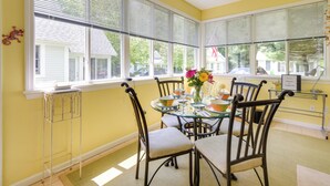 A dining area with a glass table surrounded by chairs, a vase with flowers, and a view of the outside through large windows. - Sunny Wells Cottage - 1 Mi to Moody Beach! (Wells)