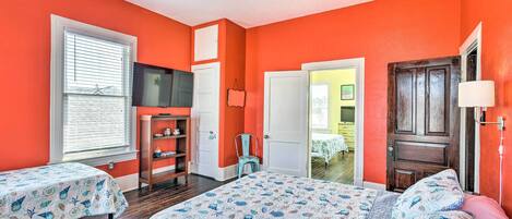 A bedroom with two beds, a ceiling fan, a window with blinds, a wooden door, and a small cabinet.