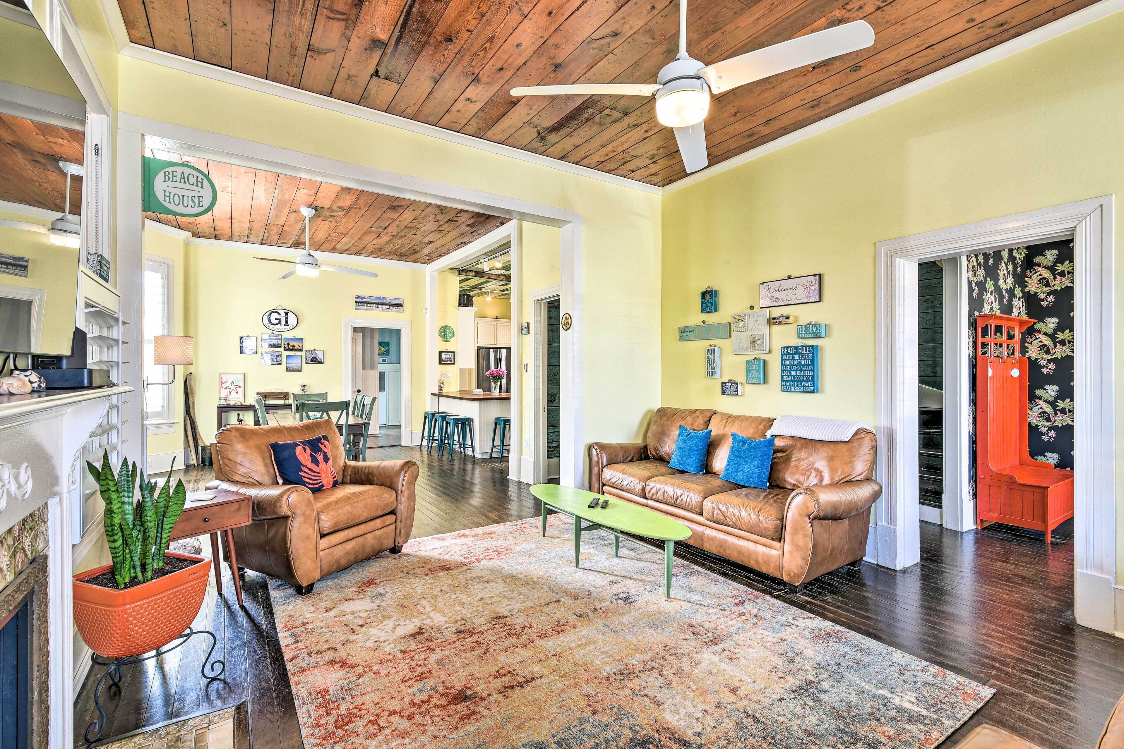 A spacious living room with a brown leather sofa, a green coffee table, and a patterned rug. A ceiling fan and a sign reading "BEACH HOUSE" are visible.