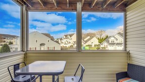 A covered patio with a table and chairs, a wicker sofa, and a view of a residential neighborhood through large windows.