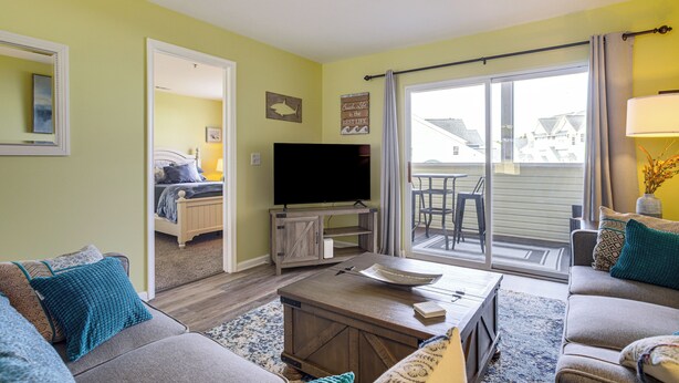A living room with a sofa, coffee table, and a flat-screen TV. There is a sliding glass door leading to a balcony.