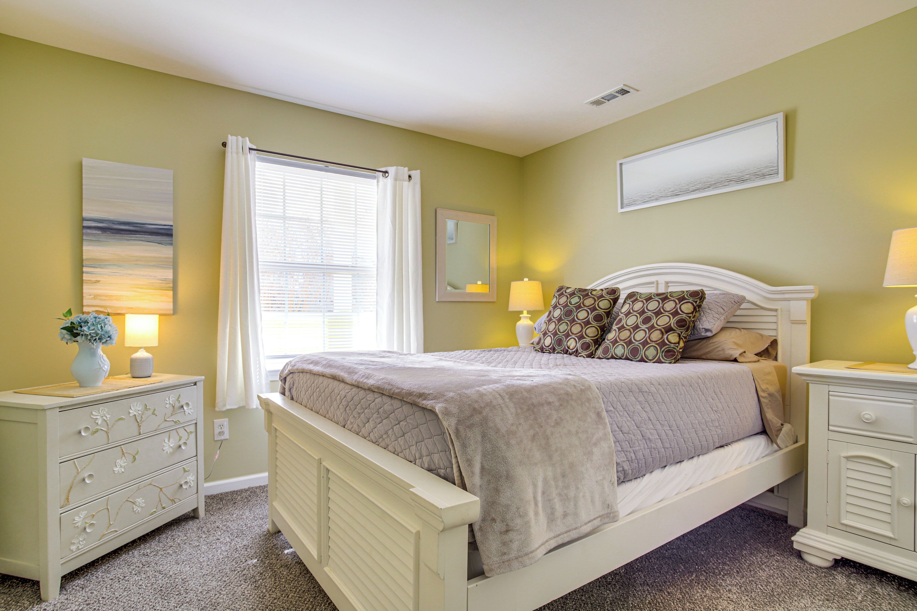 Une chambre à coucher comprenant un lit, deux tables de chevet, une commode, une fenêtre avec des rideaux et un miroir fixé au mur.