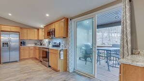 A modern kitchen with wooden cabinets, a stainless steel refrigerator, and a microwave. There is a sliding glass door leading to an outdoor deck with a table and chairs. - Hike, Swim & Ski: Home w/ Deck in Lackawaxen (Lackawaxen)