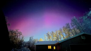 Ein Haus mit verschneitem Einfahrt und Garage, umgeben von Bäumen unter einem Nachthimmel mit sichtbarem Polarlicht.