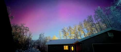A house with a snow-covered driveway and a garage, surrounded by trees under a night sky with a visible aurora.