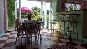 Un espace repas avec un sol à carreaux, une table entourée de chaises, un coin bar avec des tabourets et une vue sur la verdure à travers la porte vitrée.