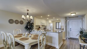 A spacious dining area with a white table and chairs, a chandelier, and a kitchen in the background. - 1501 Island Club - Folly Field - 2 Bed 2 Bath Sleeps 4 - Hilton Head Rentals (Hilton Head island)