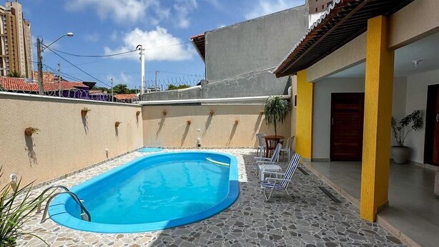 Une piscine entourée d’une terrasse dallée de pierres, un patio couvert avec des chaises, et un grand immeuble en arrière-plan.