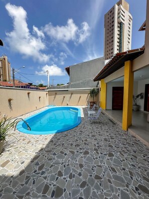 A swimming pool with a stone-tiled deck, a covered patio area with chairs, and a tall building in the background. - Hotel Pousada Aveki (Natal)