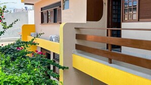 Un bâtiment moderne aux murs extérieurs jaune et beige, doté d’un balcon avec une rambarde en bois et agrémenté de plantes en pot.