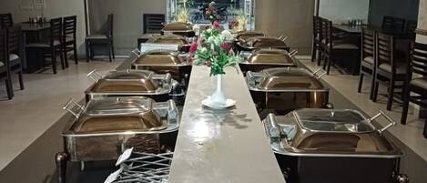 Una larga mesa de comedor con múltiples estaciones de servicio de alimentos, sillas y un florero decorativo en el centro.