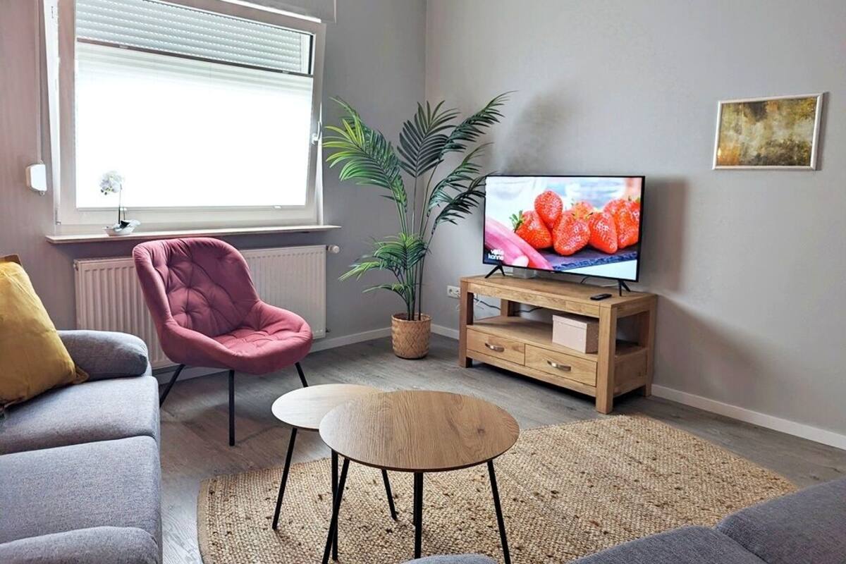 Uma sala de estar com um sofá cinzento, uma poltrona rosa, uma mesa redonda pequena, um móvel de televisão de madeira com uma TV exibindo morangos, uma planta grande em vaso e um radiador.