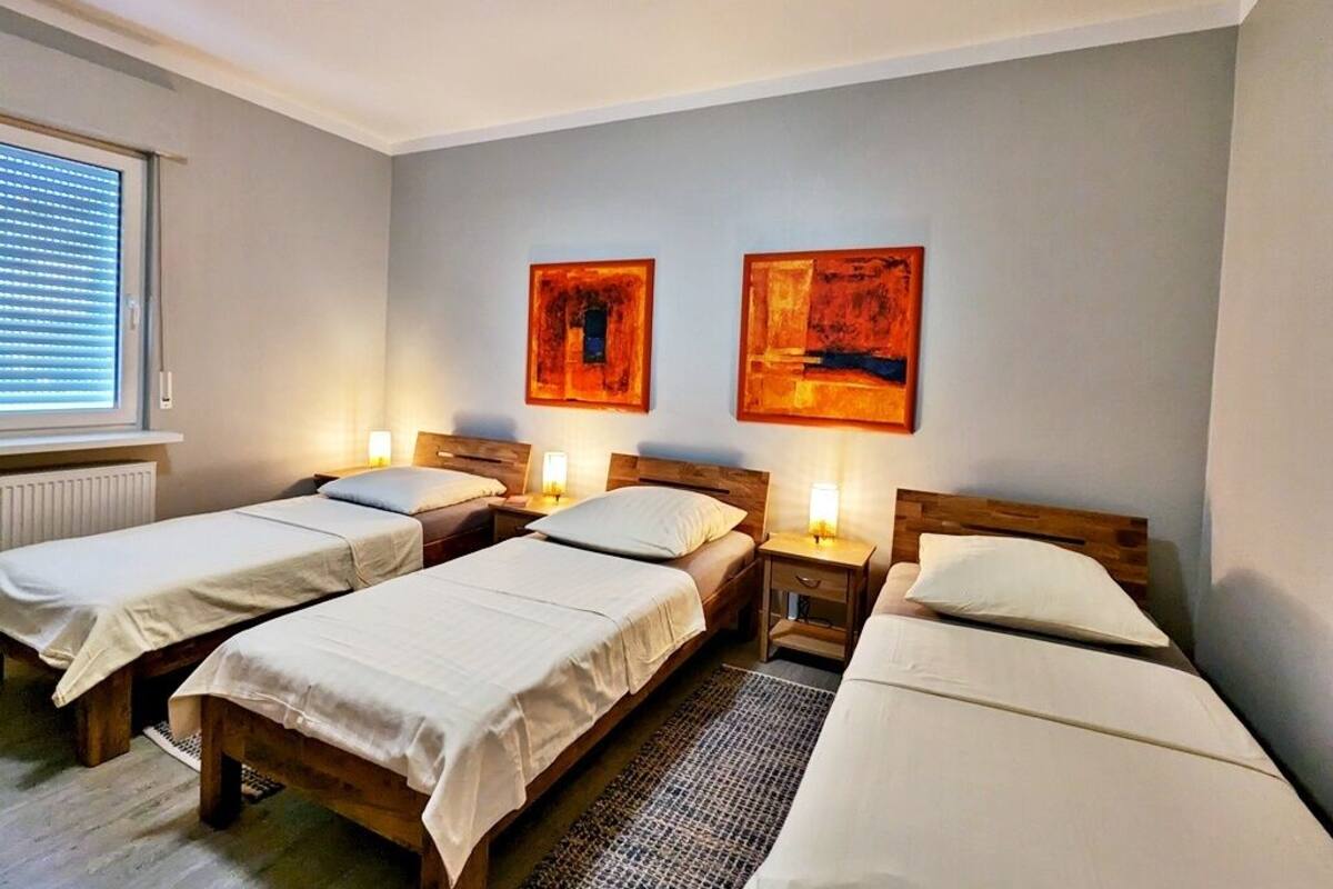 Quarto com três camas de solteiro, cada uma com cabeceira de madeira e roupa de cama branca. Há duas pinturas penduradas na parede e o quarto possui uma janela com persianas.