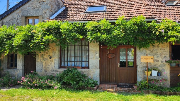 A stone building with a tiled roof, a wooden door, and a window with a curtain. There is a sign on the door and a potted plant on the right side. - La Chalaumière (Chalaux)
