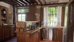 A kitchen with wooden cabinets, a built-in dishwasher, a coffee maker, and a window with curtains. - La Chalaumière (Chalaux)