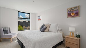 A modern bedroom with a large bed, a side table, a chair, and a window with a view of buildings. - Winton Wonder - Wanaka Holiday Apartment (Wanaka)