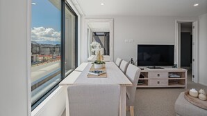 A modern living room with a large TV, a dining table, and a view of a cityscape through large windows. - Winton Wonder - Wanaka Holiday Apartment (Wanaka)