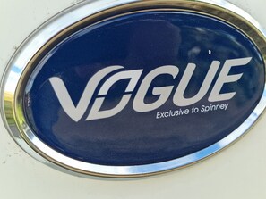 A circular emblem with the word 'Vogue' and 'Exclusive to Spinney' on a metallic surface. - Brand new Touring Caravan Sited all Setup Ready (Holyhead)