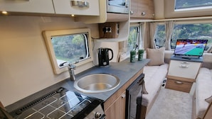 Cucina compatta per camper con fornello, lavello e un piccolo oblò che si affaccia sul verde.