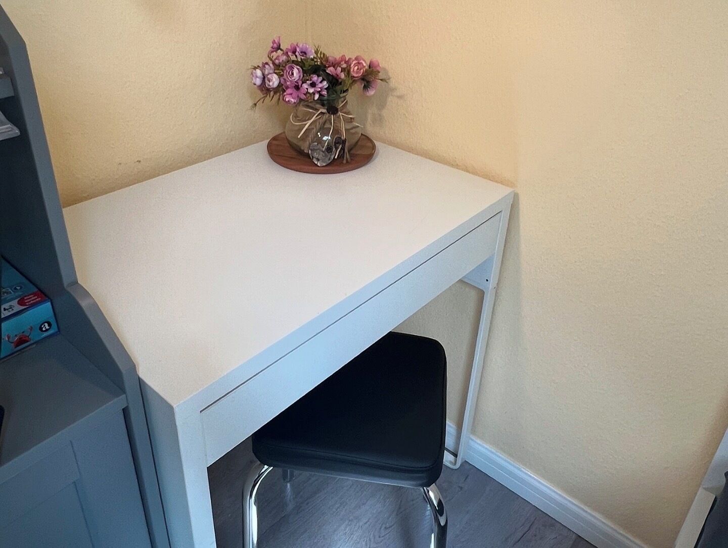 Ein kleiner, weißer Schreibtisch mit einem schwarzen Stuhl darunter. Ein Blumenstrauß in Rosa und Violett steht auf dem Schreibtisch.