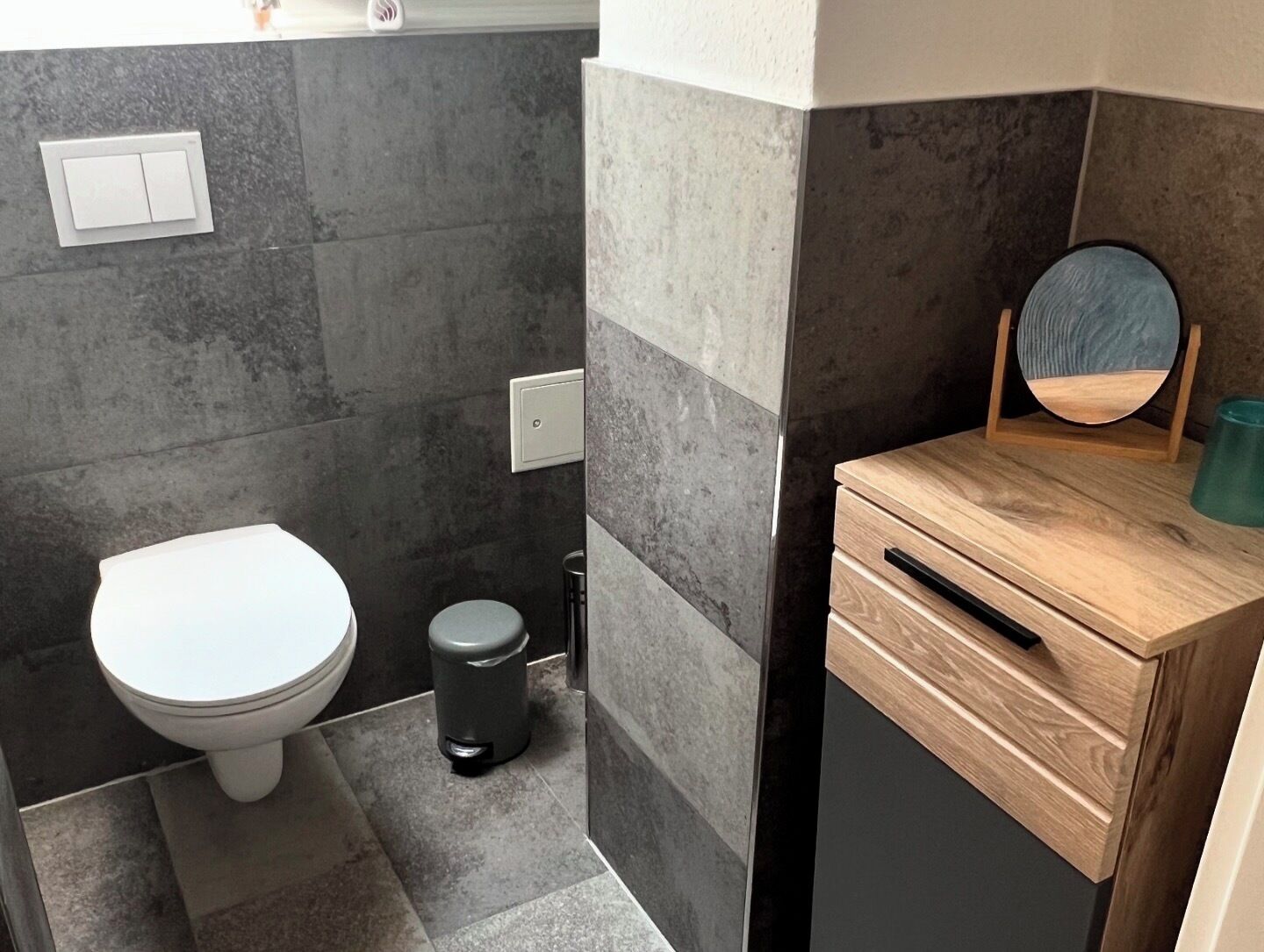 A modern bathroom with a toilet, a trash bin, and a wooden vanity with a mirror.