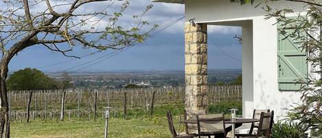 A rustic outdoor seating area with a table and chairs, a stone pillar, and a view of a vineyard and distant landscape.