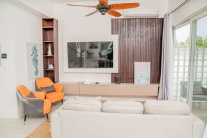 Un salon moderne avec un canapé, des fauteuils orange, un ventilateur de plafond, un téléviseur et un panneau mural en bois.