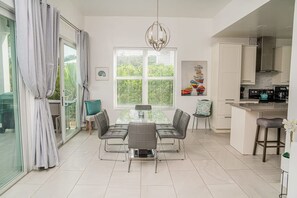 Une cuisine moderne avec des meubles blancs, un coin repas doté d’une table en verre et une grande fenêtre avec des stores.