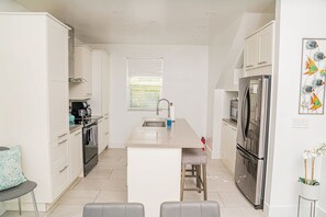 Une cuisine moderne avec des meubles blancs, un îlot central, des appareils en acier inoxydable et une fenêtre équipée de stores.