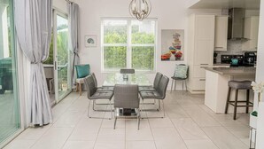 Una cucina moderna con mobili bianchi, un'area pranzo con tavolo in vetro e un'ampia finestra con persiane.