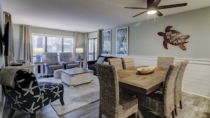 A cozy living room with a dining area, featuring wicker furniture, a ceiling fan, and artwork on the walls.