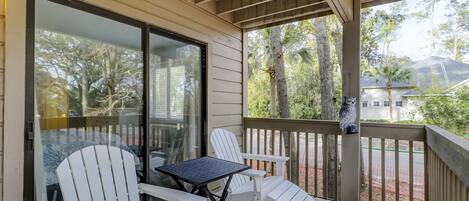 A covered porch with white Adirondack chairs and a small table, overlooking a residential area with trees and houses.