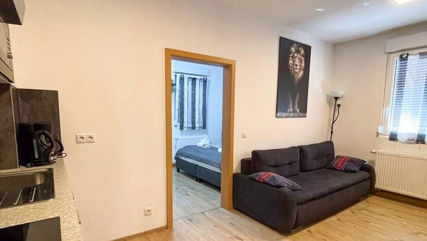 Uma sala de estar moderna com sofá, mesa de centro e televisão. Há uma porta que dá para um quarto com cama e janela.
