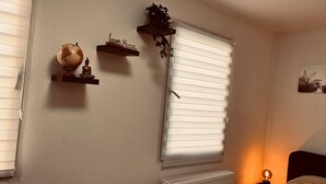 Quarto com cama, radiador, globo terrestre, prateleira com estátua de Buda, janela com persianas e planta suspensa na parede.