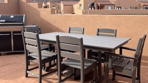 Een terras buiten met een barbecue, tafel en stoelen, met uitzicht op een bergachtig landschap.