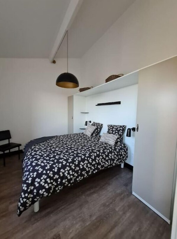 A bedroom with a bed, a bench, a hanging light fixture, and a built-in shelf.