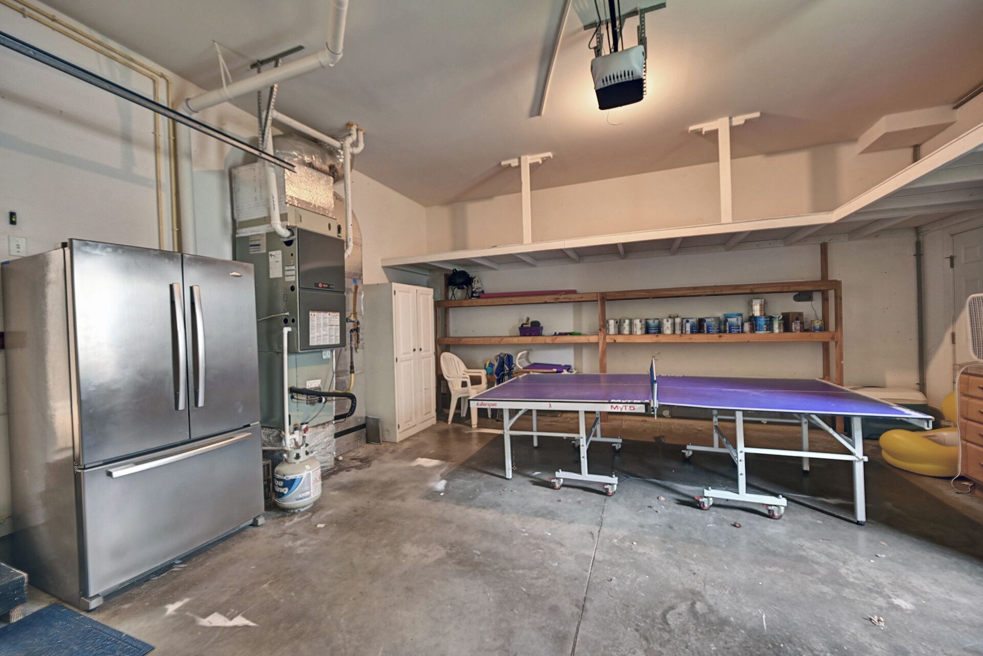 A compact kitchen with a stainless steel refrigerator, a gas cylinder, and a ping pong table.