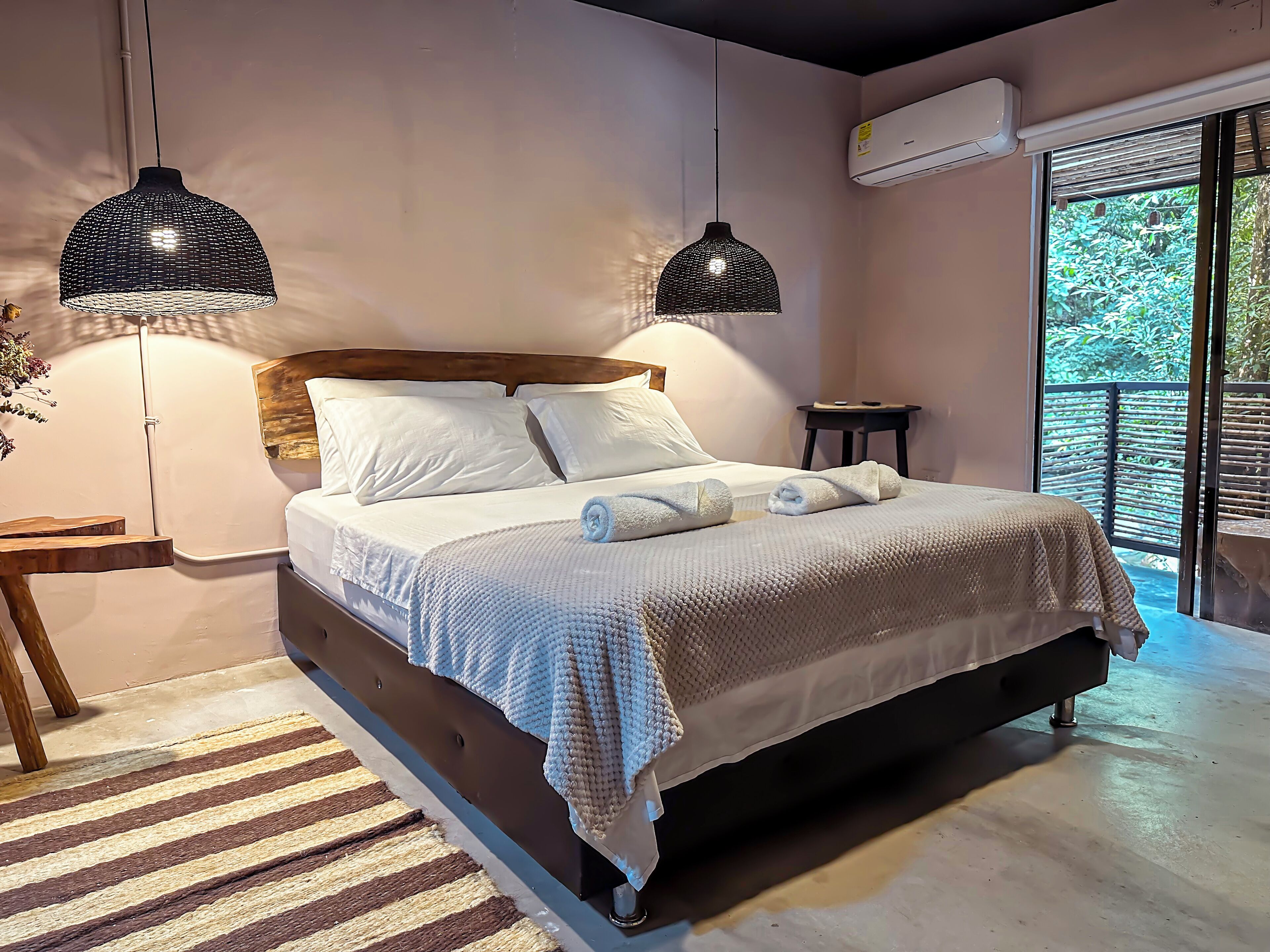 A bedroom with a bed, two pendant lights, a nightstand, a small table, and a view of greenery outside.