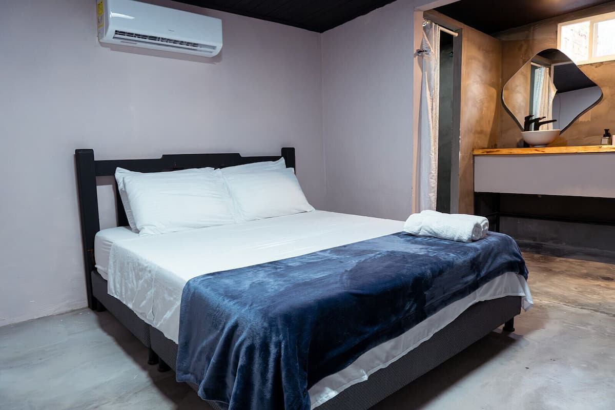 A single bed with white linens and a blue blanket, a wooden headboard, a white air conditioning unit above, and a wooden vanity with a mirror and a bowl.