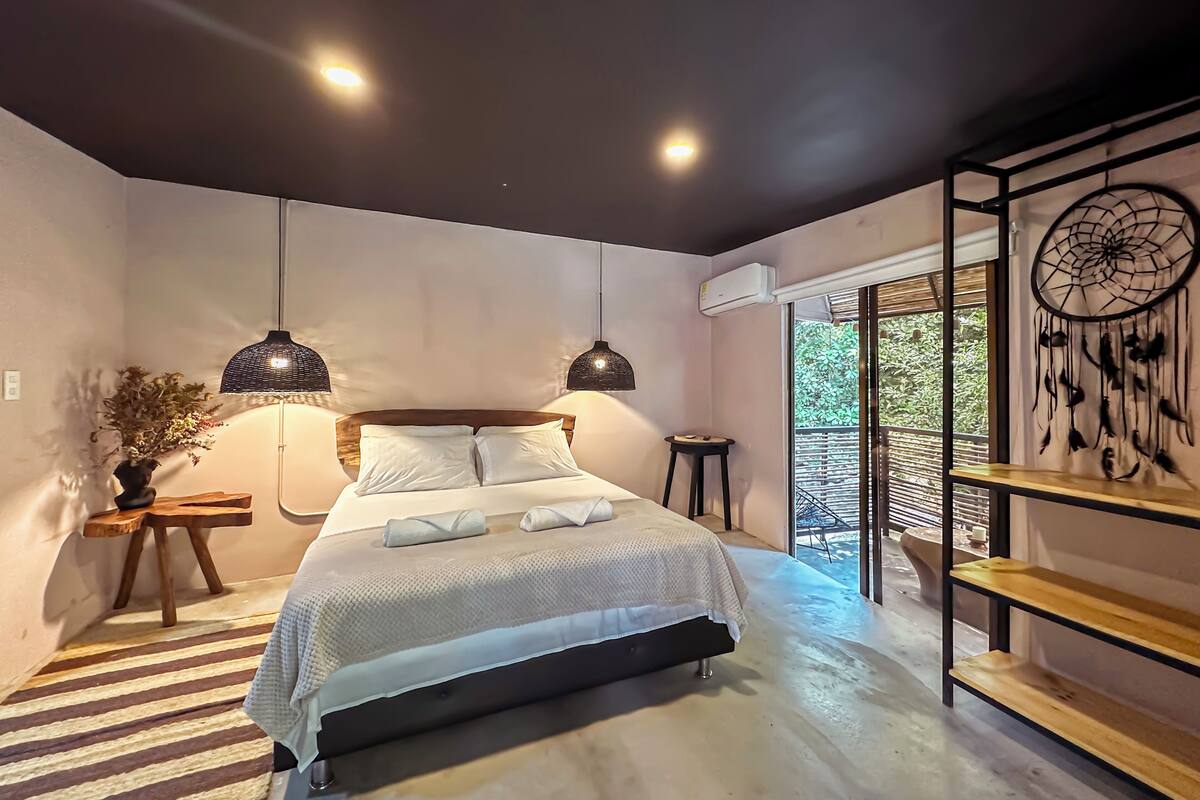 A modern bedroom with a large bed, two pendant lights, a dreamcatcher, and a view of greenery outside.
