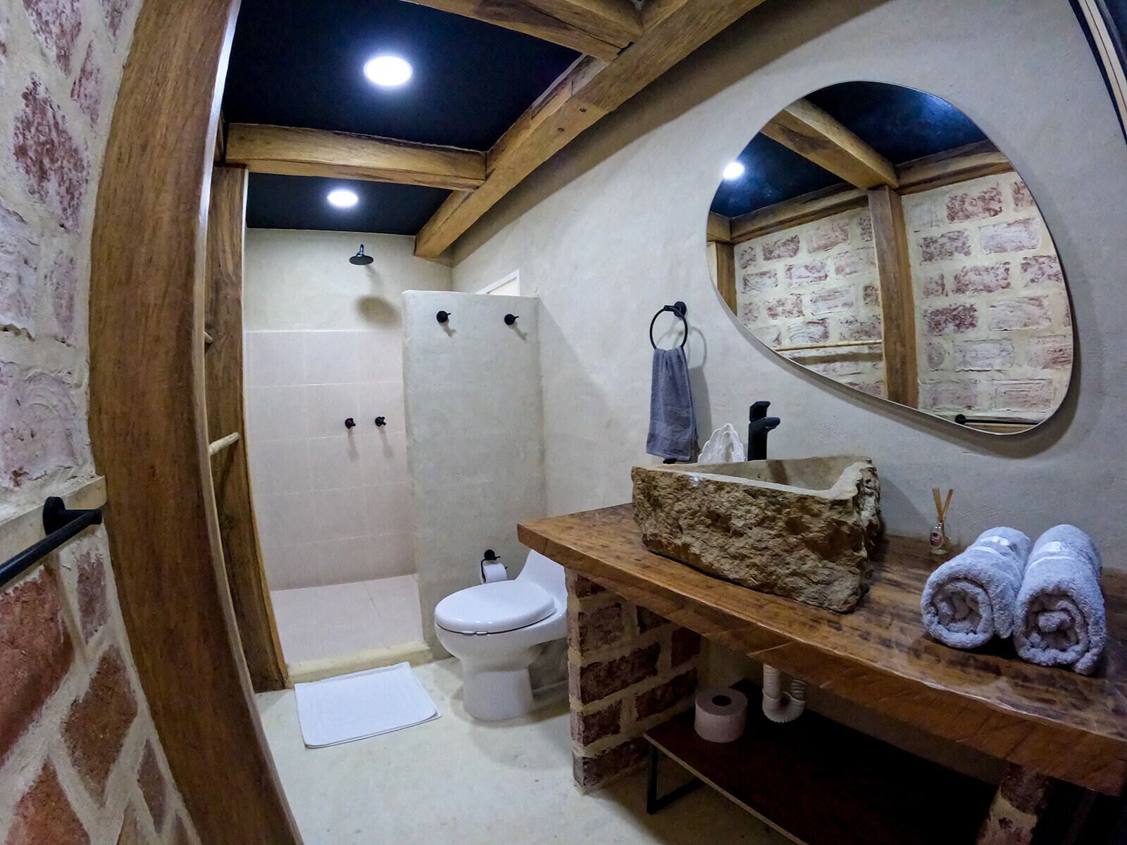 A bathroom with a wooden vanity, a white toilet, a shower, and a mirror with a brick wall design.
