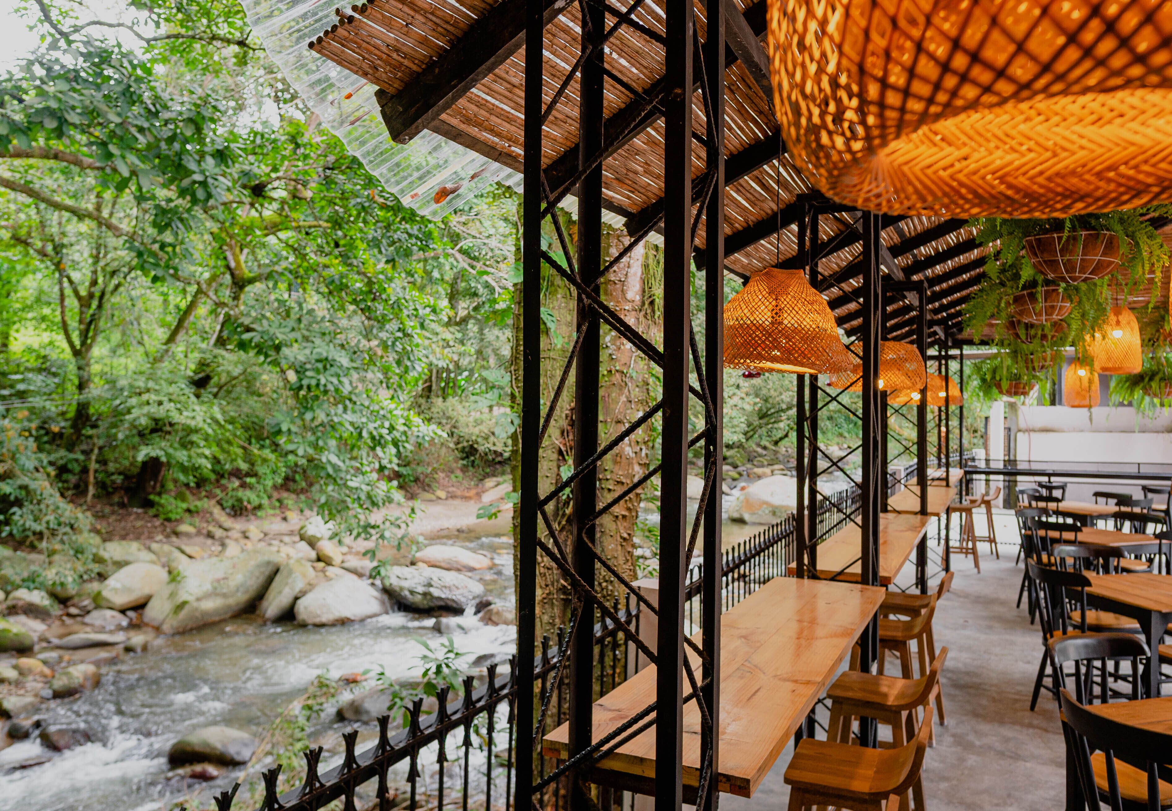 Un restaurante al aire libre con mesas y sillas de madera, en un entorno natural con un arroyo y árboles.