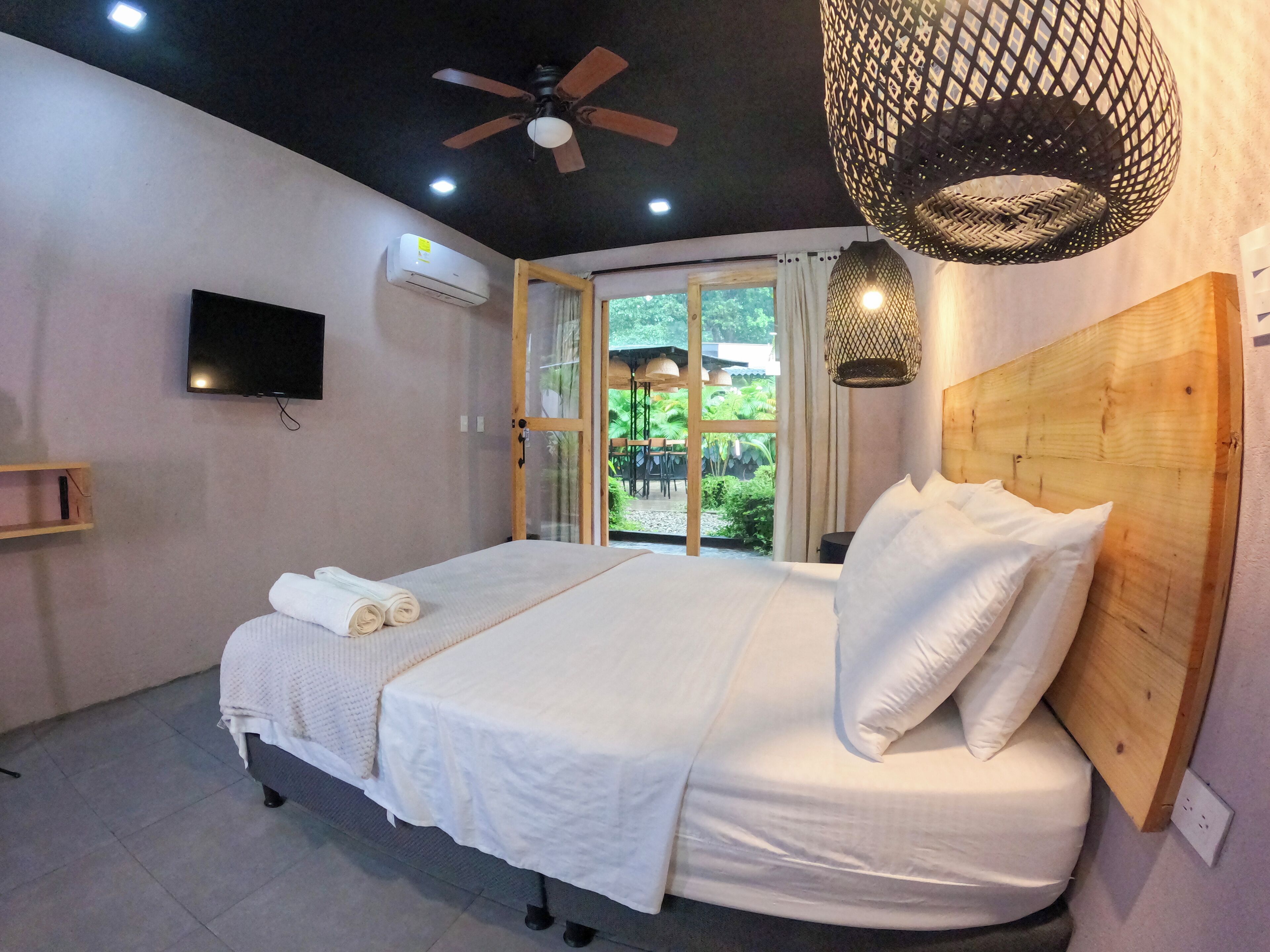 Un dormitorio con cama, ventilador de techo, televisor y ventana con vista a la vegetación.