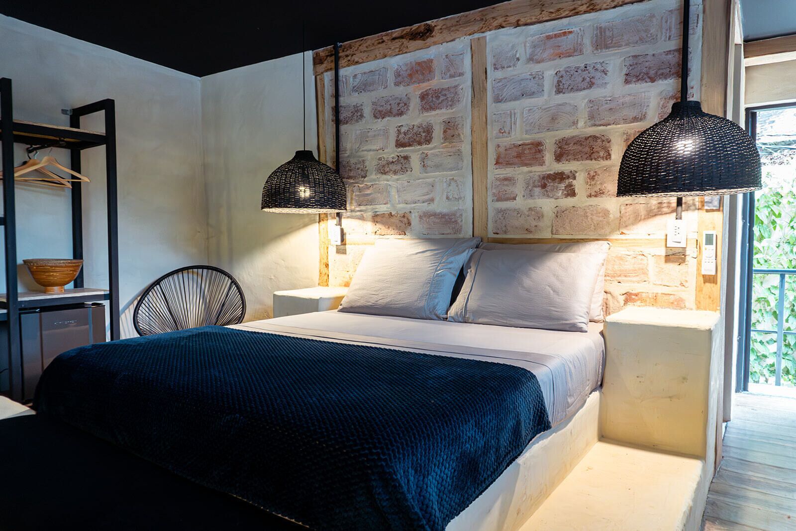 A bedroom with a brick wall, a bed with white linens, two black pendant lights, a wooden chair, and a small shelf with a bowl.