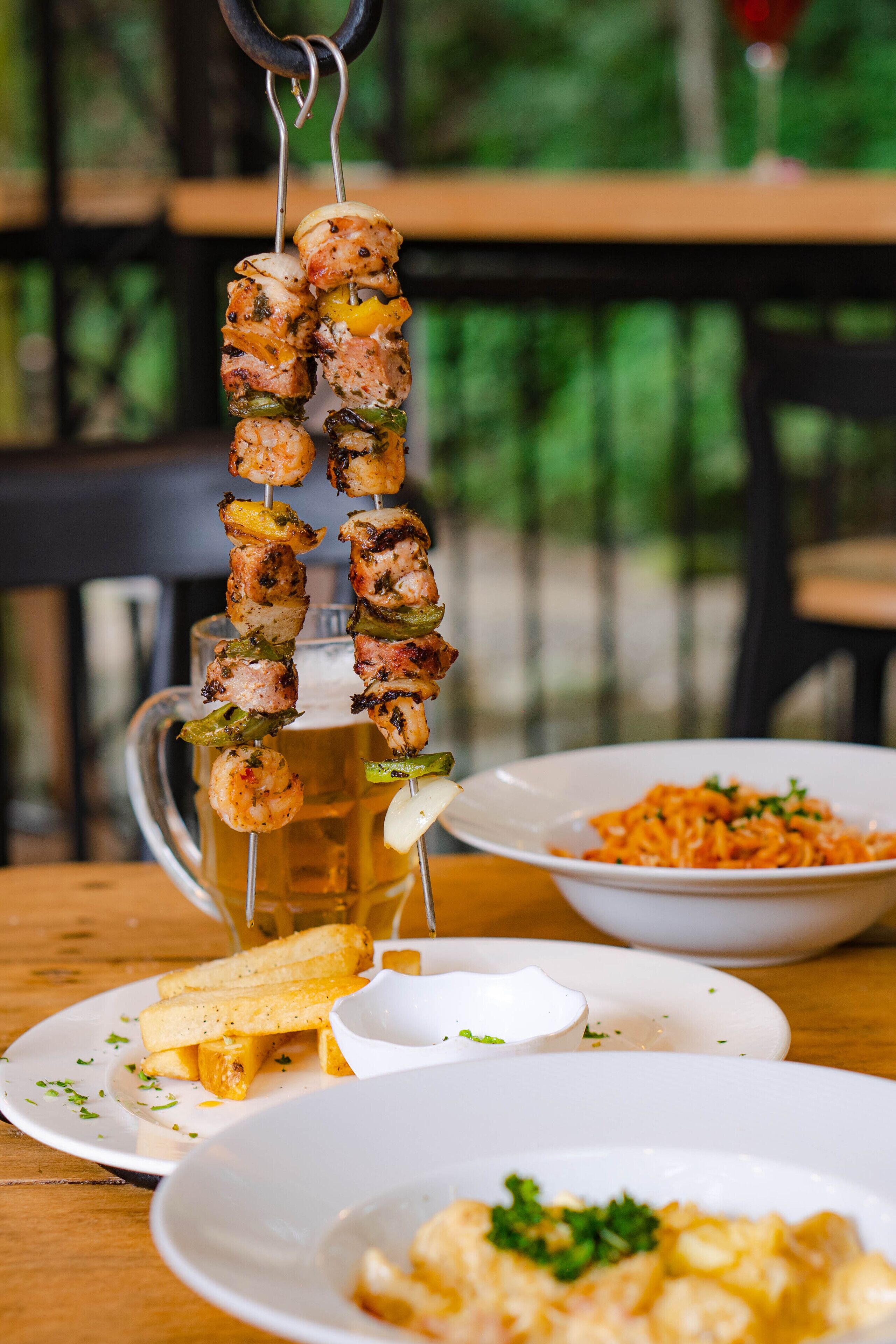 A meal setup with skewers of grilled meat and vegetables, a beer mug, and plates of pasta and fries.