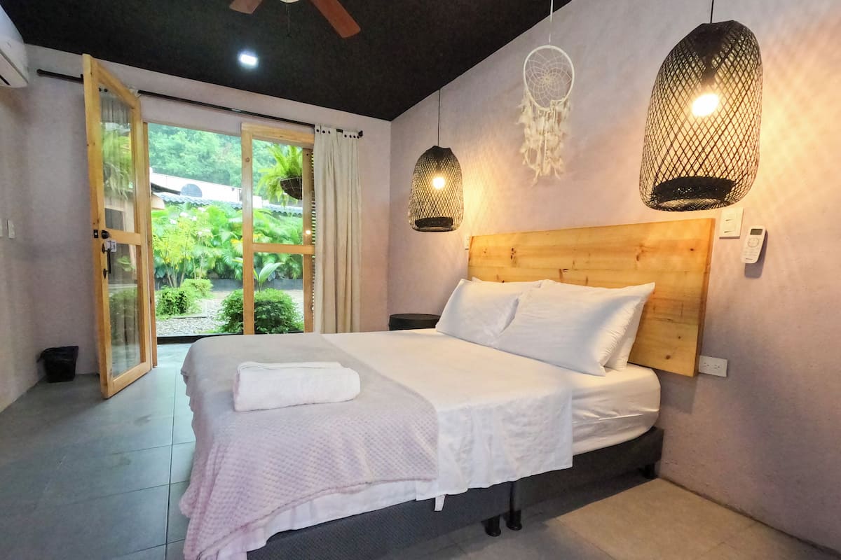A bedroom with a wooden headboard, a bed with white linens, a nightstand, a dreamcatcher hanging from the ceiling, and a view of greenery outside.