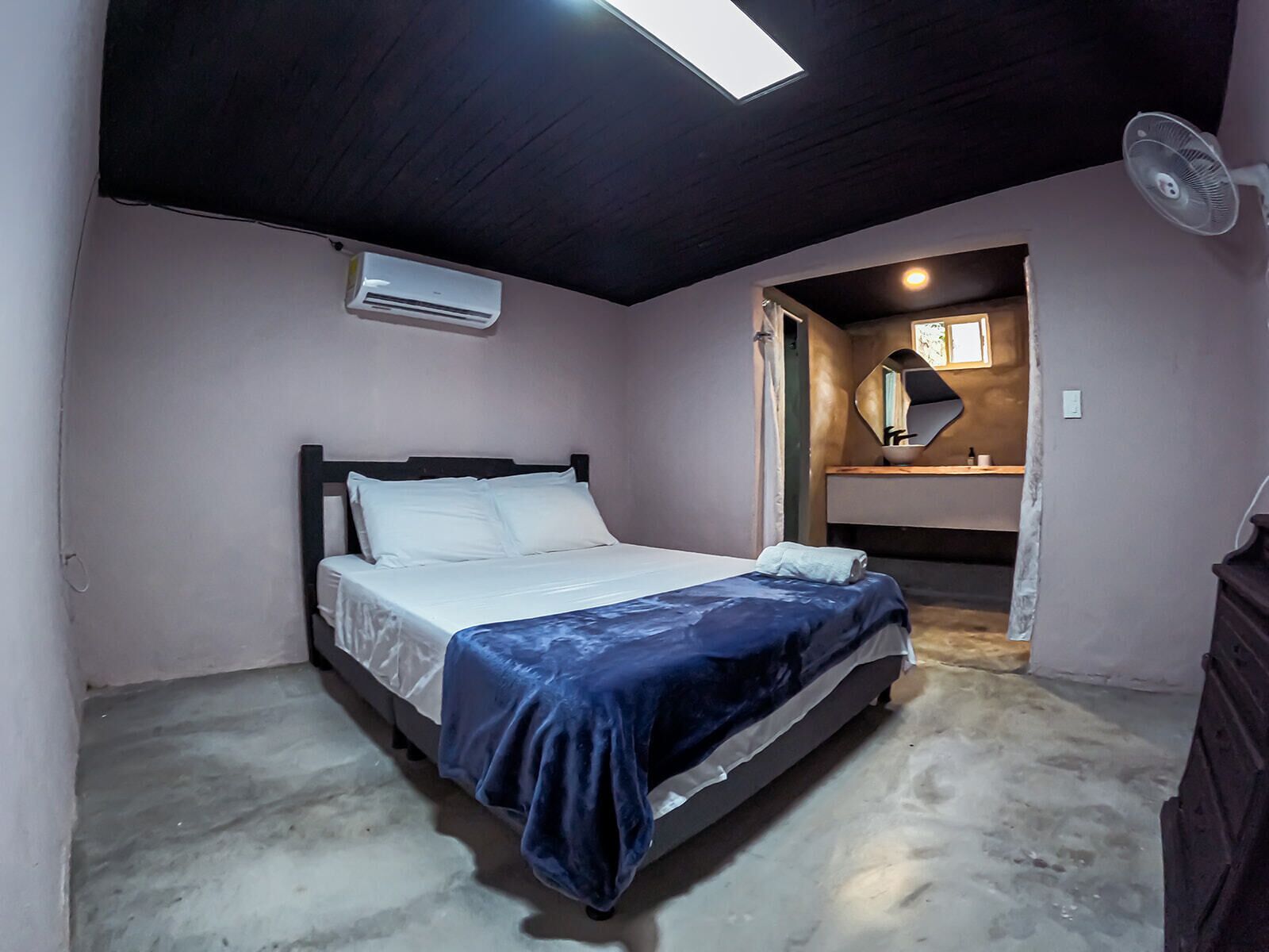 Un dormitorio con cama, aire acondicionado, ventilador de techo, espejo y baño pequeño.