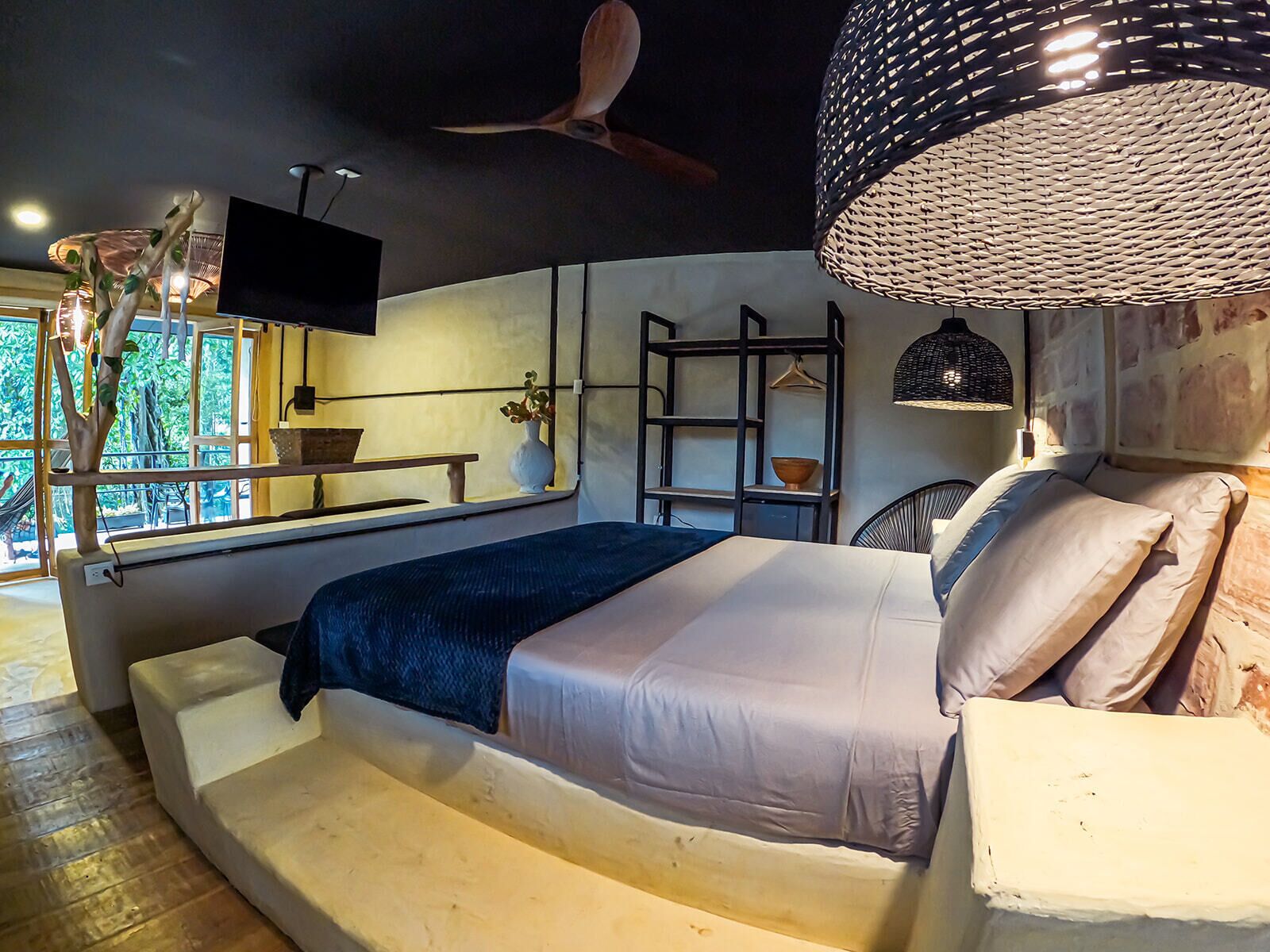 A modern bedroom with a bed, a ceiling fan, a hanging light fixture, a window with a view of greenery, and a small table with a vase.