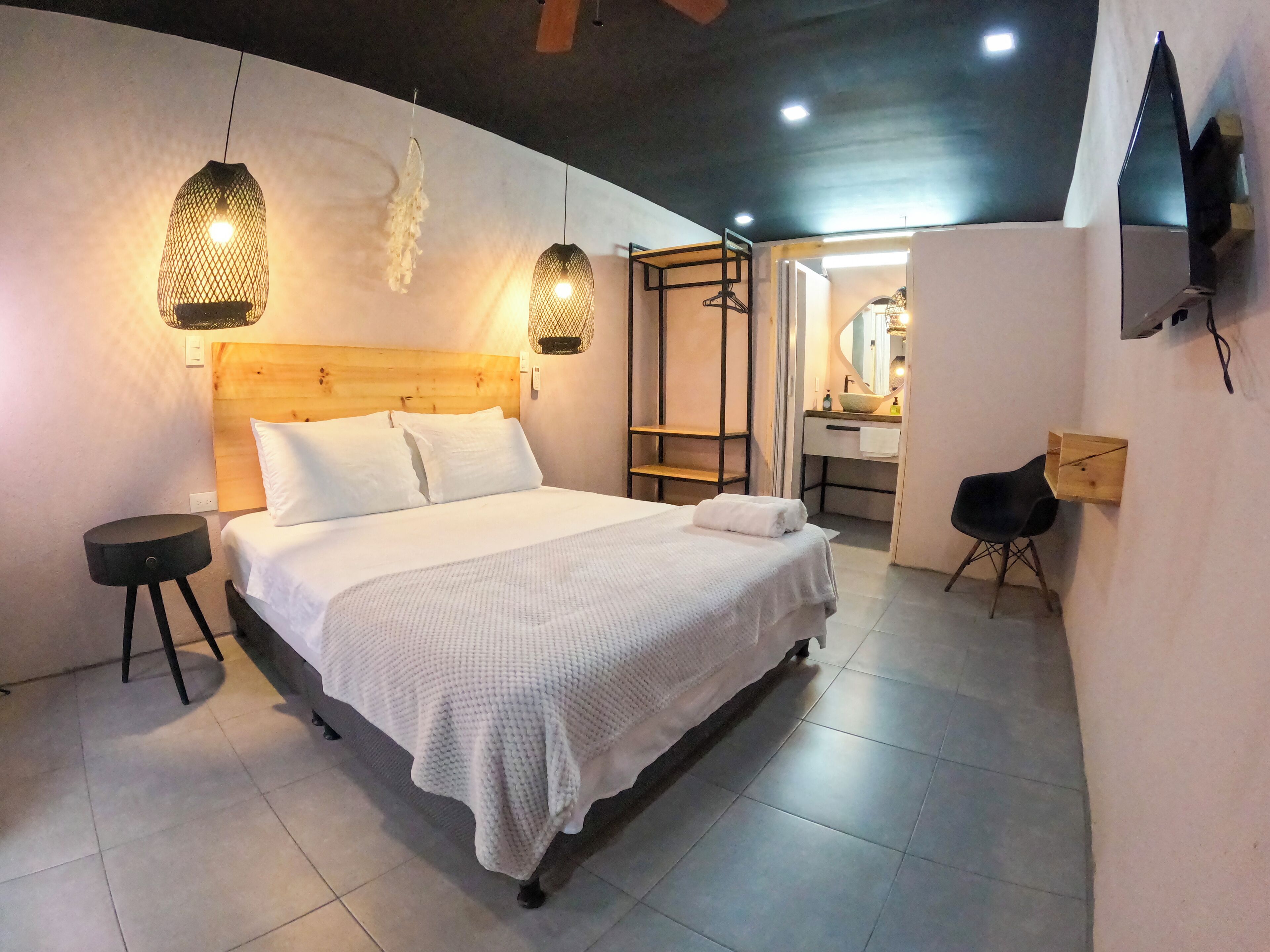 Un dormitorio moderno con cabecera de madera, cama con ropa de cama blanca, dos lámparas colgantes, una mesita de noche redonda negra, una silla de madera y un baño con espejo y lavamanos.
