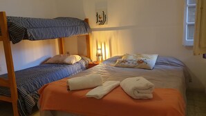 A room with two beds, one with a bunk and one with a single mattress. There are pillows and a blanket on the beds, and a lamp on the nightstand.